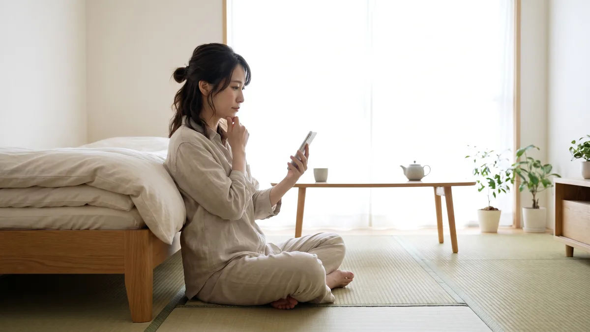 たたんだ布団のそばでスマートフォンを見ながら料金を考える女性（イメージ）